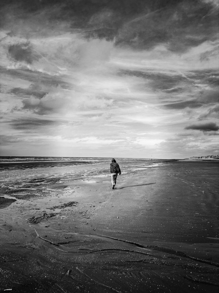 Black and white photo of a person walking alone along a wide, empty beach under a dramatic cloudy sky